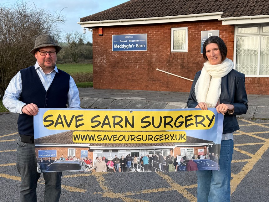 Carl Peters-Bond with Meddygfa'r Sarn campaign organiser, Clare Treharne in front of the surgery holding a protest banner